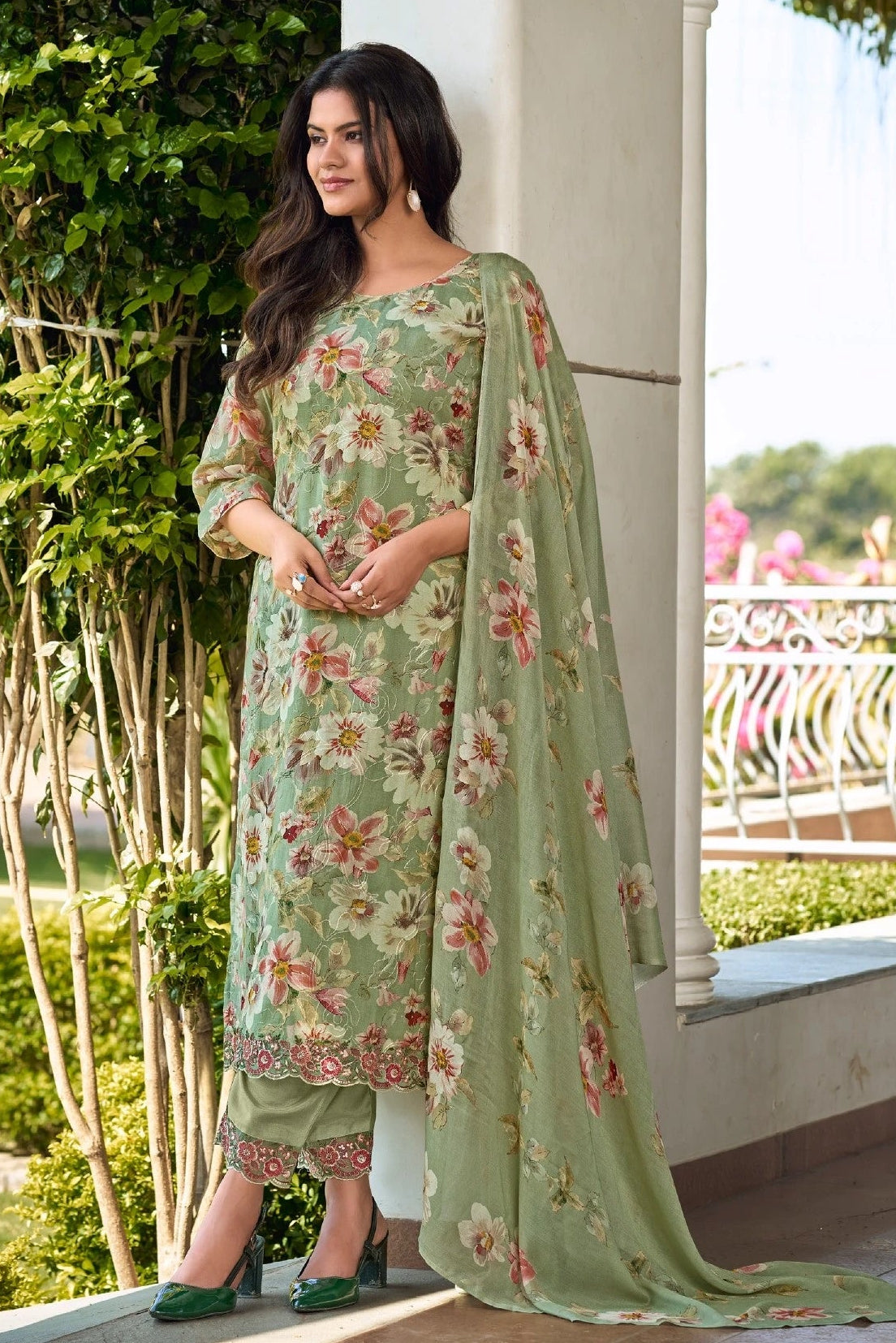 Woman in a floral dress standing outdoors with greenery and a white building in the background